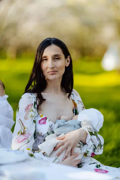 Güzel bir parkta, bir kadın sıcak mevsimde doğanın canlı renkleriyle çevrili yavrusunu şefkatle emziriyor.