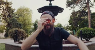 Huzurlu bir park, güneş gözlüklü sakallı bir adamı bir çeşmenin yanında lotusstil otururken gösteriyor, doğanın sakinliğini ve akan suyu düşünürken.