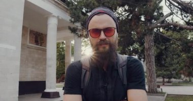 Sakallı ve güneş gözlüklü kendine güvenen bir adam modern bir binanın önünde duruyor, kollarını kavuşturmuş, şehir ortamında kararlılık ve otorite yayıyor.