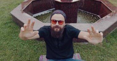 Sakallı ve güneş gözlüklü rahat bir adam resimli bir fıskiyenin önündeki çimlerin üzerinde huzurlu bir anın tadını çıkarıyor.