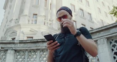 Sakallı bir adam şehir manzarasını geziyor, telefonuyla meşgul. Güneş gözlüğü takıyor, mesajları sırt çantasıyla kontrol ediyor, modern teknolojiyi gösteriyor.