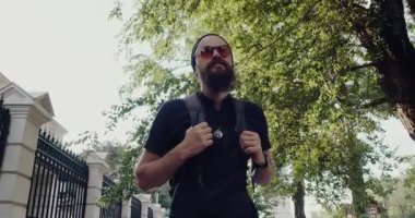 Güneş gözlüklü sakallı bir adam sırt çantasıyla şehir caddesinde yürüyor. Bir ağacın yanında duruyor, çevresinin tadını çıkarıyor, şehir keşiflerini somutlaştırıyor.