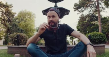 Sakallı bir adam, sakin bir parkta, otların üzerinde otururken, sakin ve sakin bir şekilde, huzurlu bir ortamda, içsel bir gözlem yapıyor.