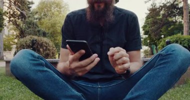 Bu videoda dışarıda şık sakallı bir adam var. Telefonunu kullanıyor ve kulaklık takıyor. Modern şehir yaşamını yansıtan bir çeşmenin yanında boş vakitlerin tadını çıkarıyor.