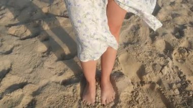 Beyaz elbiseli, kumlu bir sahilde çıplak ayakla duran bir kadın. Dalgalar ayaklarına kapanır, sakin bir atmosfer yaratır ve doğal güzelliklerini sergiler.