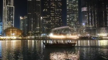 Karanlık gökyüzüne bakan hareketli bir şehrin sakin bir gece manzarası. Suda yüzen bir tekne, şehir gece hayatını gözler önüne seriyor.