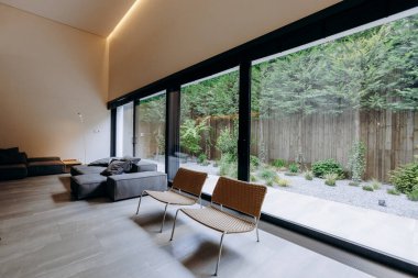 Geniş oturma odasında geniş pencerelerin bolluğu, geniş doğal ışığın yanı sıra birkaç konforlu sandalye bulunmakta.