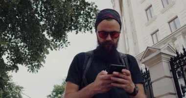Şık güneş gözlüklü şık bir genç adam dışarıda dolaşırken akıllı telefonunu kullanıyor.