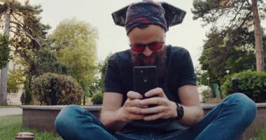 Sakallı bir adam otların üzerinde oturuyor, cep telefonuna dalmış. Bu yalnızlık anı, doğanın ortasında teknolojiye odaklanmasına zıt düşüyor.