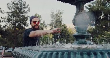 Güneş gözlüklü sakallı bir adam güneşli bir parkın tadını çıkarır. Güzel bir çeşmeye hayran kalır ve su ile etkileşime geçer.