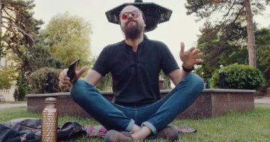 Sakallı bir adam parkta çimlerin üzerinde nilüfer bir poz içinde oturuyor, cep telefonuna dalmış, huzuru modern teknolojiyle harmanlıyor.