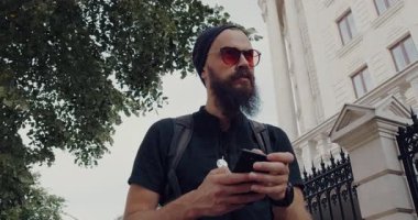 Sakallı ve güneş gözlüklü şık bir adam, modern bir binanın önünde mesajları kontrol etmek için durmadan önce, telefonuyla meşgul bir şekilde caddede yürüyor.