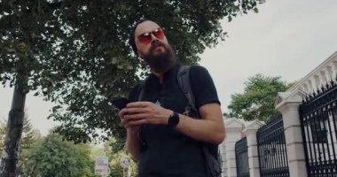 Sakallı bir adam şehir merkezinde geziniyor, yürürken telefonunu kontrol ediyor. Bir ağacın yanında duruyor, sonra güneş gözlüğü takıyor, cihazına odaklanıyor.