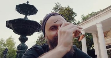 Huzurlu bir parkta, güneş gözlüklü sakallı bir adam büyüleyici bir çeşmenin yanında, doğada huzurlu ve rahat bir atmosfer sergiliyor.