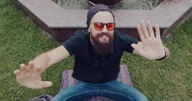 Güneş gözlüklü sakallı bir adam çeşmenin yanındaki çimlere oturur, kollarını açar, güneşin tadını çıkarır ve akan suyun rahatlatıcı sesinin tadını çıkarır.