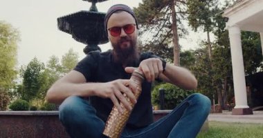 Açık havada sakin bir ortamda sakallı bir adam çeşmenin kenarında çimlerin üzerinde otururken neşeli bir şekilde davul çalar, su içer ve bir sandviçin tadını çıkarır.