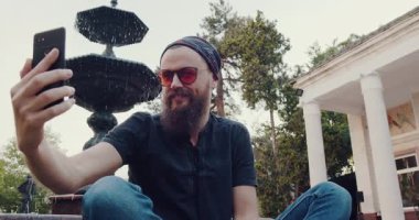 Sakallı bir adam güzel bir fıskiyenin yanında poz vermekten ve eğlenceli selfie çekmekten hoşlanır. Sahne rahatlık ve neşe saçıyor.