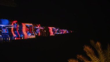 Canlı bir şehrin ufuk çizgisi en yüksek aydınlatılmış bina, yanıp sönen ışıklar ve sallanan bir palmiye ağacına ev sahipliği yapar ve canlı şehir atmosferini yükseltir.