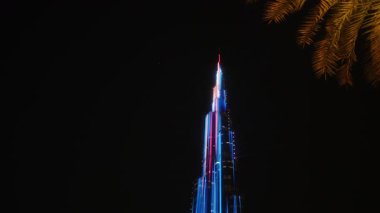 Burj Halife 'nin muhteşem gece manzarasına tanık olun, en yüksek bina, mavi ve kırmızıyla aydınlatılmış, palmiye ağaçları güzelliğini artırıyor.