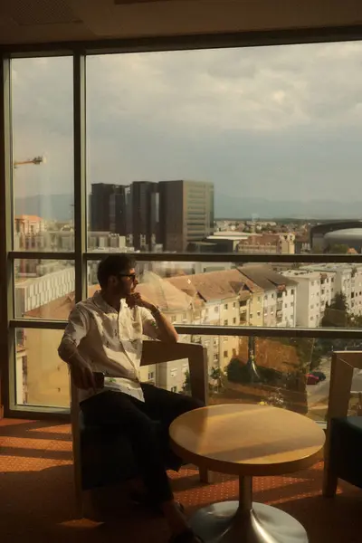 Bir adam büyük bir pencerenin önünde rahatça oturuyor, hayat ve aktivite dolu hareketli bir şehre bakıyor.