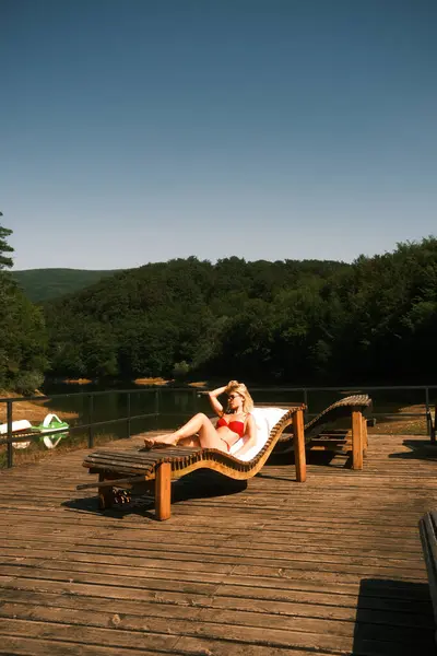 Canlı kırmızı bir bikini giyen bir kadın su kenarındaki ahşap iskelede rahat bir şezlongda dinleniyor.