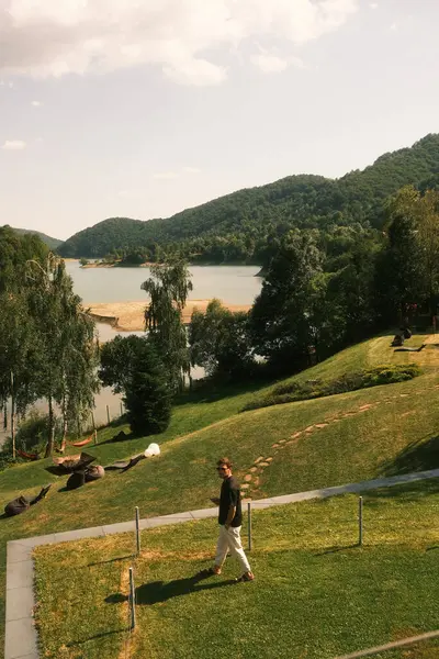 Bir adam sakin bir gölün arka planda gökyüzünü güzel bir şekilde yansıttığı pitoresk bir parkta rahatça yürüyor.