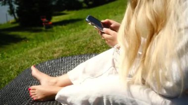 Sakin bir açık hava ortamında, beyaz cüppeli bir kadın bir battaniyenin üzerinde rahatlıyor, cep telefonuyla sükuneti birleştiriyor.