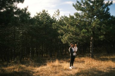 A couple stands closely together in a tranquil forest environment, surrounded by swaying trees and golden grass