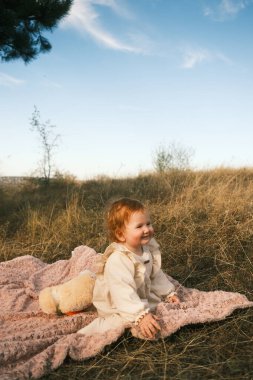A cheerful toddler is joyfully enjoying the beautiful outdoors, sitting on a cozy blanket with a teddy bear