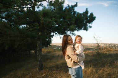 A loving mother holds her cheerful smiling child outdoors, surrounded by lush trees and the beauty of nature