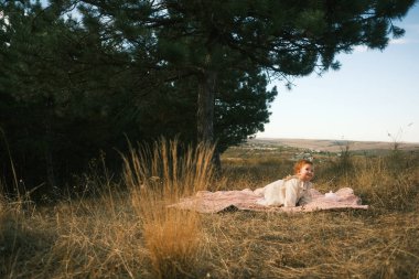 A tranquil scene of a person relaxing on a warm blanket in a grassy field, surrounded by tall, lush trees