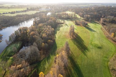 Havadan güzel bir sonbahar sabahının panoramik manzarası, golf sahası, nehir, çok renkli ağaçlar ve yeşil çimenler, sıcak güneş ışığı, gölgeler.