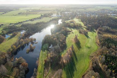 Havadan güzel bir sonbahar sabahının panoramik manzarası, golf sahası, nehir, çok renkli ağaçlar ve yeşil çimenler, sıcak güneş ışığı, gölgeler.
