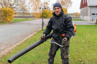 İsveç, Knislinge 26 Ekim 2022: Bir adam büyük bir hava üfleme makinesiyle düşen sonbahar yapraklarını temizliyor