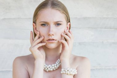 Mavi gözlü, doğal saçlı ve makyajlı, beyaz ahşap arka planda inci kolye ve bilezik takan güzel bir kızın portresi. Manken elleriyle yüzüne dokunarak poz veriyor. Boşluğu kopyala.