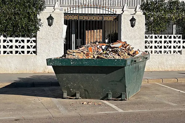 Konut binasının önünde inşaat atığı ve molozlarla dolu aşırı dolu yeşil çöp bidonu.