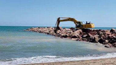 Valencia, İspanya - 08 Nisan 2025: Mavi deniz yakınlarındaki kayalık kıyı şeridi inşaatında çalışan sarı kazıcı, kıyı dalgakıranlarına kaya yerleştirdi 