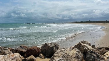Hafif dalgaların dalgalandığı kumlu sahil manzarası, ön planda büyük kayalar ve dramatik bulutlu gökyüzü ile çevrili, Costa de Valencia, İspanya