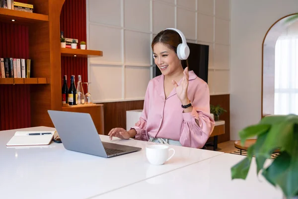 Woman in casual clothes is sitting comfortably at desk at home and wearing headphones while having a business video call to discuss, make planes, and strategize.