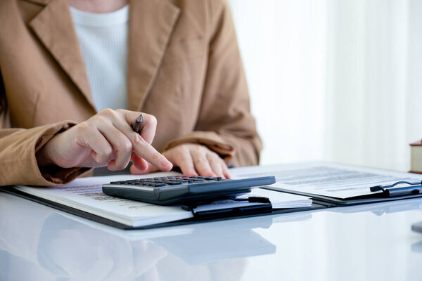 Агент по недвижимости в костюме показывает цифры и расчетный дом, стоимость недвижимости на калькуляторе для покупателя или клиента, сидя за столом с документами и моделью дома, чтобы обсудить и договориться о контракте в офисе.