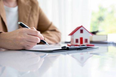 Resmi giyinmiş bir kadın, bir evi, mülkü ya da konut sigortasını masasında oturan bir ev modeliyle görüyor ve kontrol ediyor. Anlaşmayı imzalamadan önce kararını veriyor..