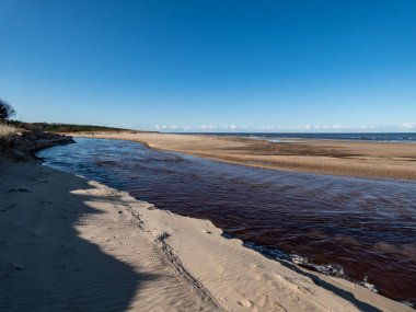 Deniz manzarası Baltık denizi ve plajını bembeyaz sarı kum ve önünde bir nehir ve yukarıda parlak mavi gökyüzü ile tasvir ediyor.
