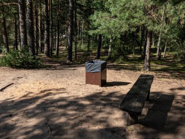 View of a trash bin and wooden bench surrounded with sand in the middle of a forest in summer