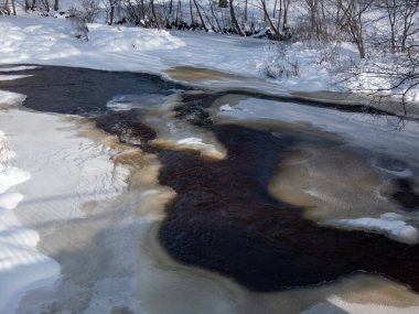 View of a big river partly frozen with visible flowing, dark water and banks and surroundings covered with white snow in winter