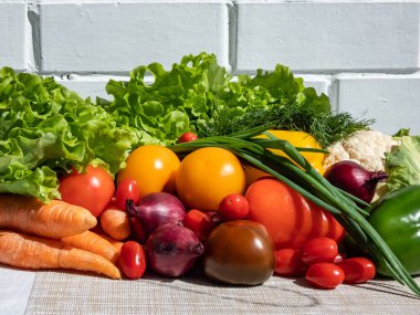 Bir yığın taze, sağlıklı ve renkli sebzeler parlak güneş ışığı altında ve arka planda beyaz tuğla bir duvar var. Domates, soğan, havuç, biber, karnabahar, salata, dereotu ve yeşiller masada.