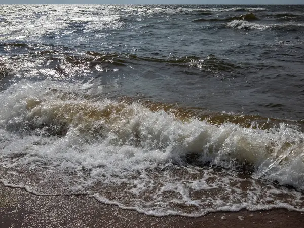 Baltık Denizi 'nin rüzgarlı bir gününde kıyıda kahverengi kum ve yukarıda mavi gökyüzü olan büyük köpüklü dalgaların yakın çekimi. Baltık Denizi 'ni tasvir eden Deniz Burnu