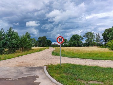 Kırmızı daire içinde 30 numaralı metal yol ya da trafik levhası yeni hız limitini gösteriyor. Taşra manzarası, mavi gökyüzü ve arka planda bulutlarla çevrili bir çakıl yolu.