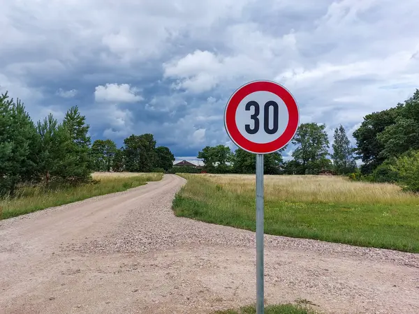 Metal road or traffic sign with number 30 in a red circle indicating new speed limit a gravel road surrounded with countryside scenery against blue sky and clouds on background
