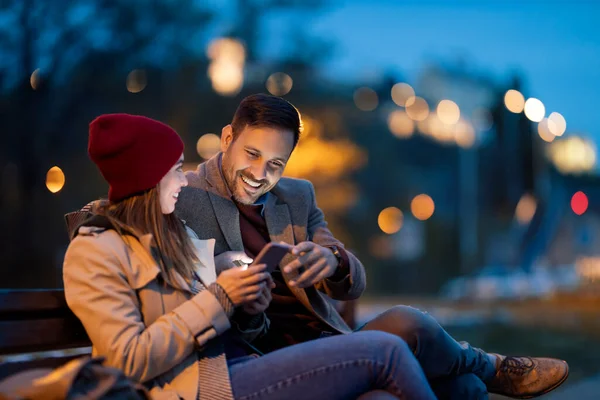 Happy young couple sitting in a park bench and using smart phone. Enjoying at night.