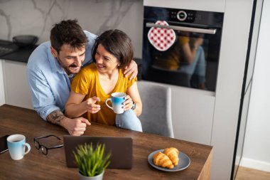 Evde otururken, kadın ve erkeğin birlikte kahve içip dizüstü bilgisayarda çalışması..